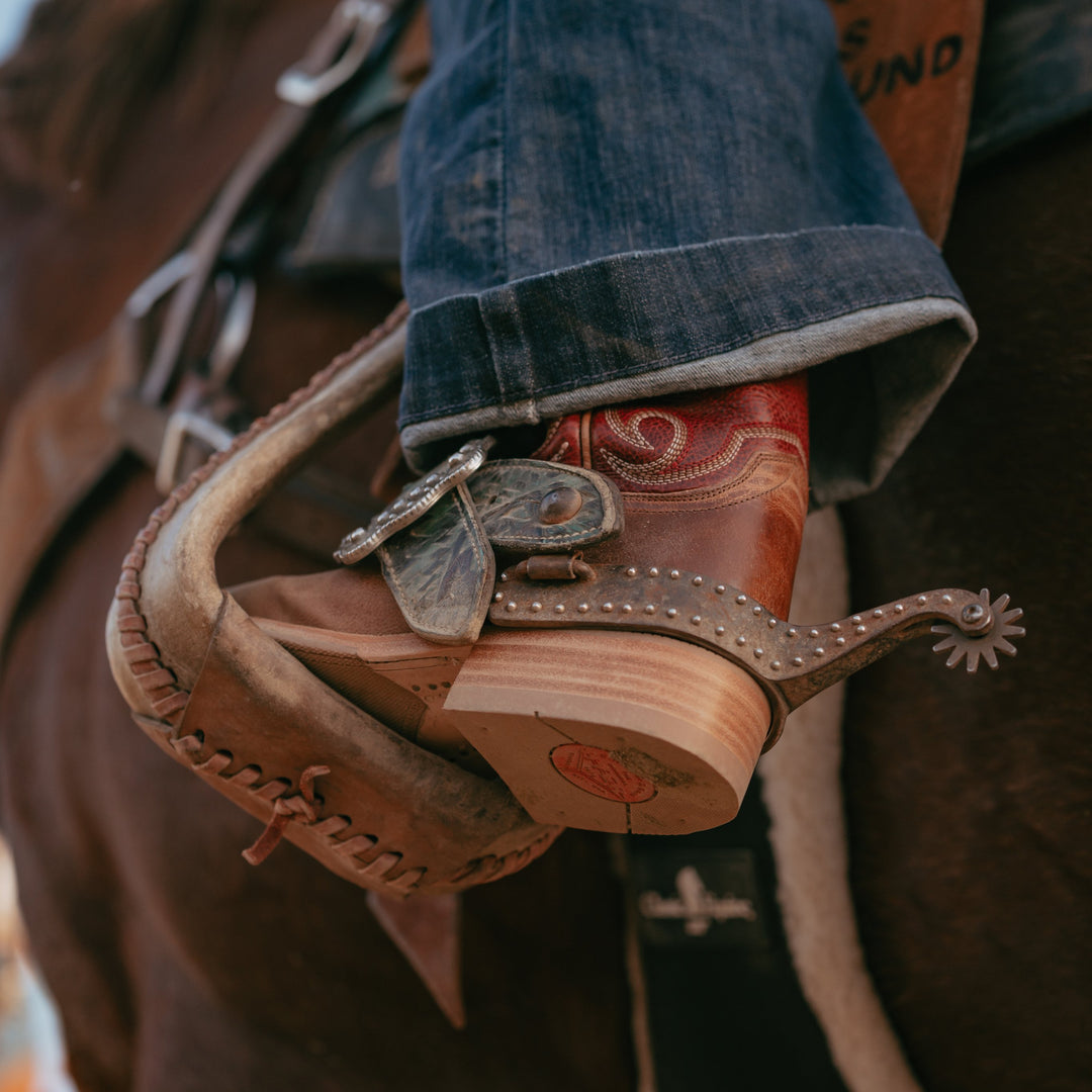 A close-up of a person's foot in a cowboy boot and cuffed blue jeans resting in a leather horse spur on a horse.