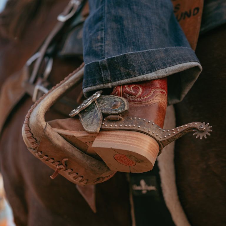 A close-up of a person's foot in a cowboy boot and cuffed blue jeans resting in a leather horse spur on a horse.