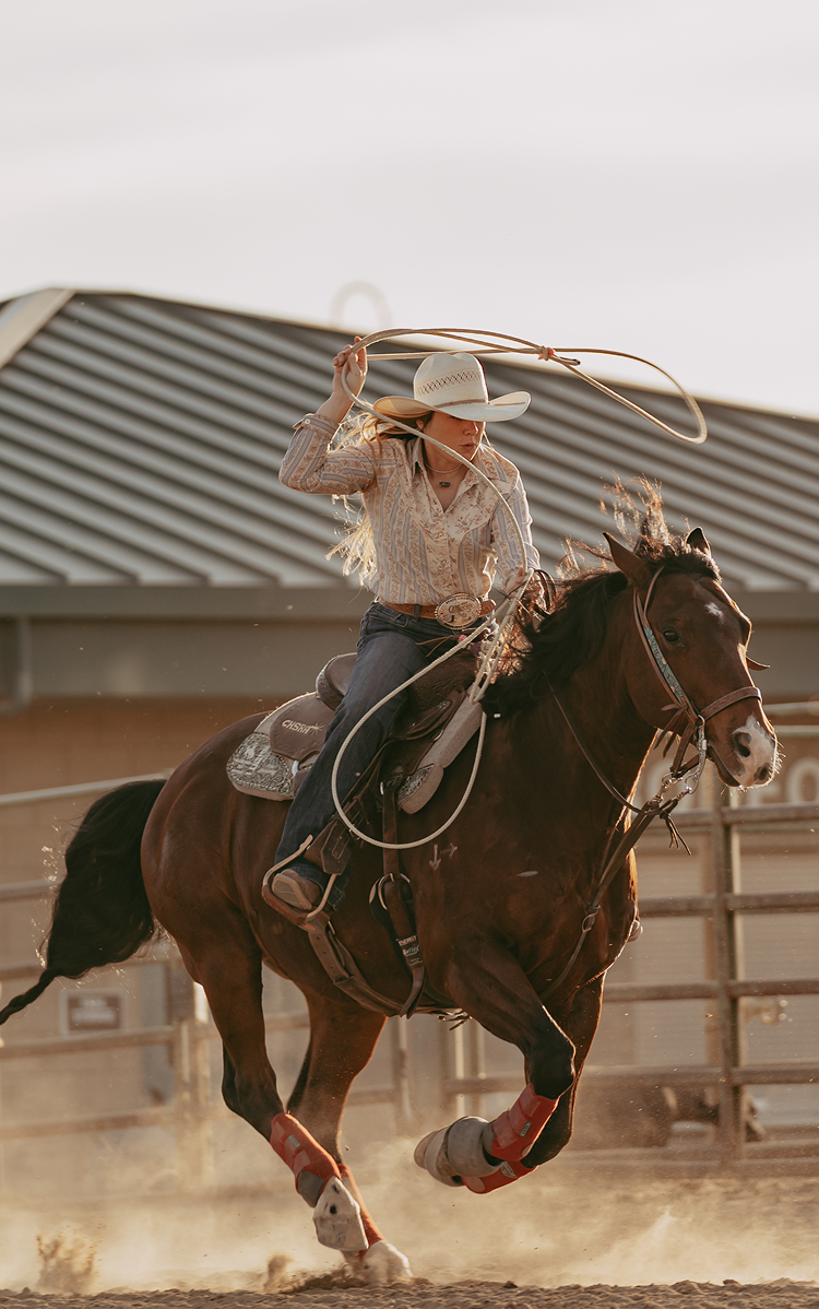 A woman in western attire rides a brown horse during a rodeo event, swinging a lasso in an outdoor arena with a metal-roofed building in the background.