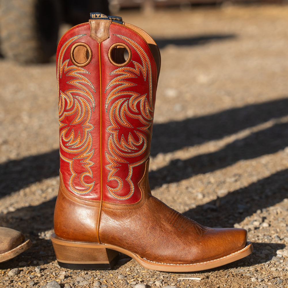 A single red and tan leather cowboy boot with ornate western embroidery on the shaft, photographed outdoors on a gravel surface in bright sunlight.
