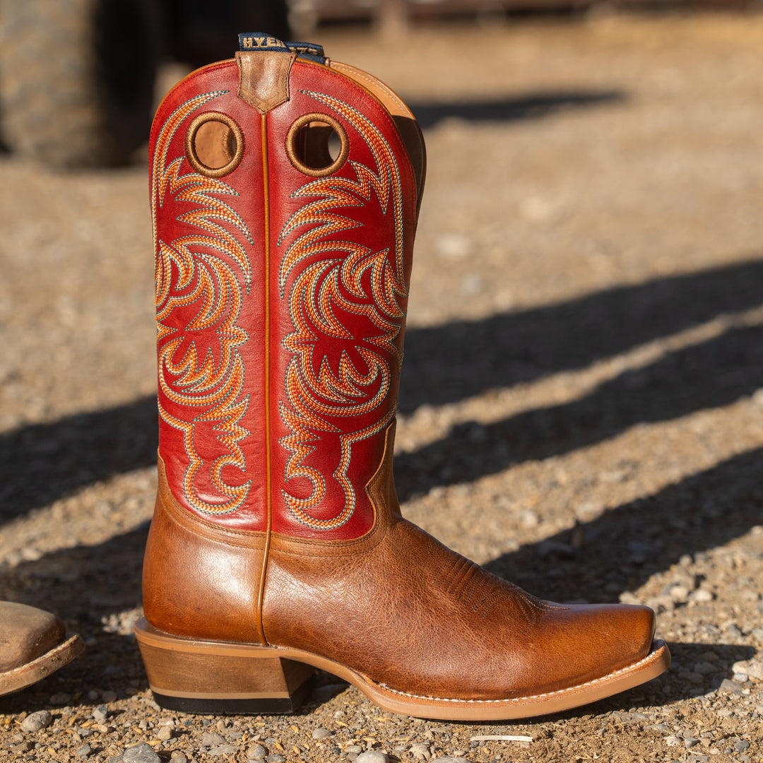 A single red and tan leather cowboy boot with ornate western embroidery on the shaft, photographed outdoors on a gravel surface in bright sunlight.
