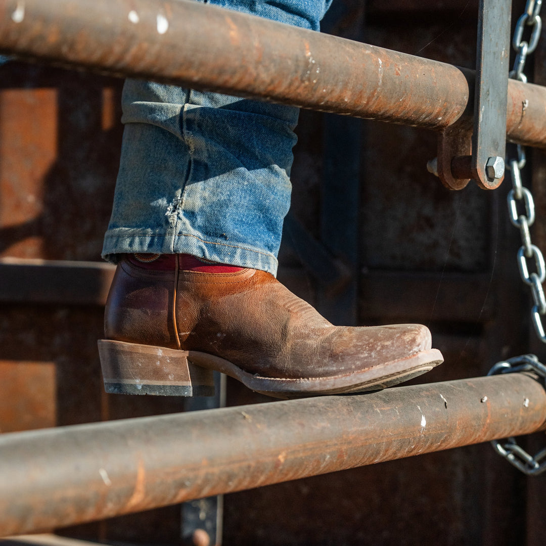 Close up photo of a person wearing a brown leather cowboy boot with their foot up on a metal fence post