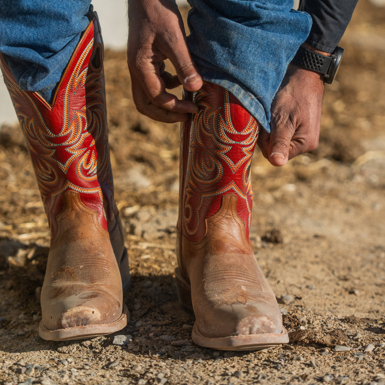 A close-up of a person's hands pulling up blue jeans on their legs, revealing a pair of tan and red leather cowboy boots with vibrant red and gold western embroidery.