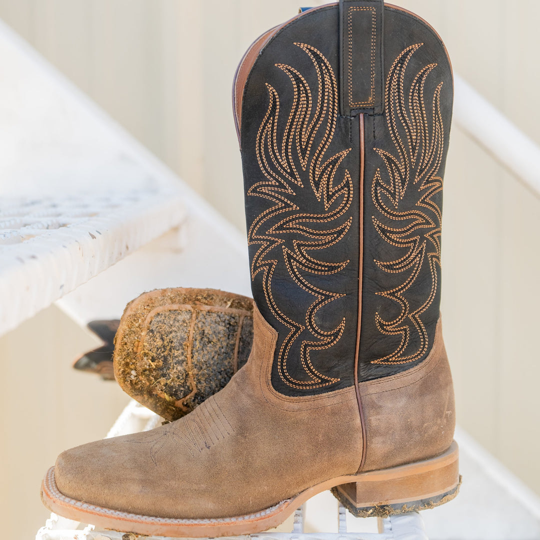 A single brown leather cowboy boot with a black shaft and tan embroidered western pattern, standing on a white surface with a blurred rock in the background.