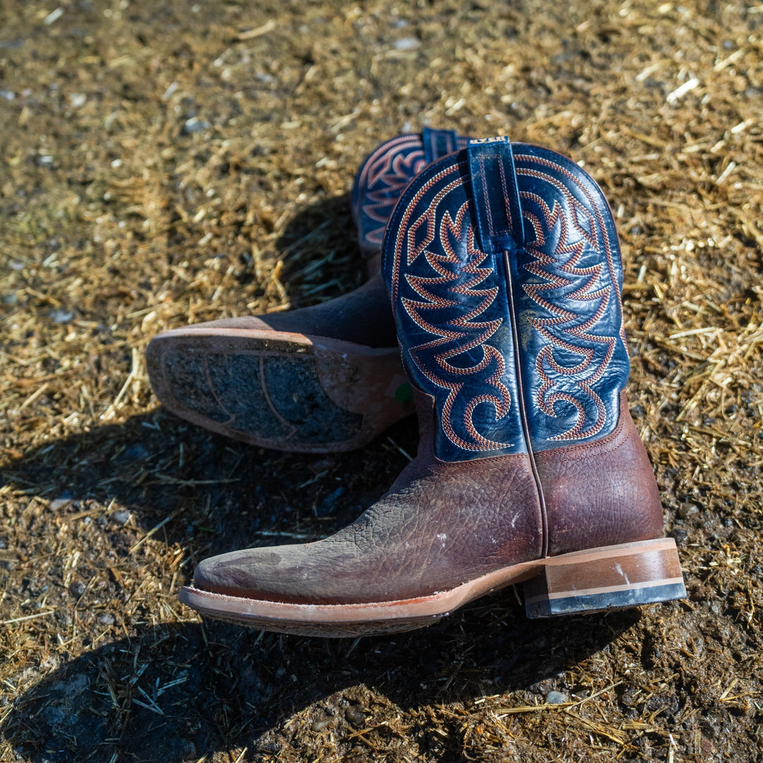 A pair of navy blue and brown cowboy boots laying on their sides on top of a muddy, straw filled background.