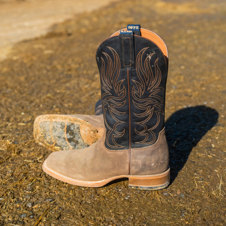 A pair of western cowboy boots standing on a dirt ground. The boots feature a brown leather lower section and a black leather upper section with tan embroidery.