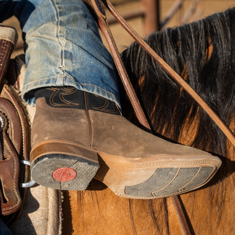 A close-up of a person's foot wearing a brown leather cowboy boot with decorative stitching, resting on a horse's footrest while wearing blue jeans.