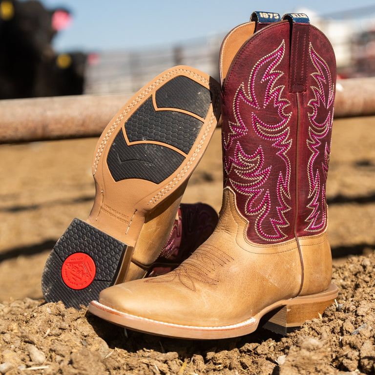 A pair of western cowboy boots standing on dirt ground, with one boot upright showing a tan leather shaft and a burgundy decorative upper, and the other boot tilted to show the black treaded sole with a red circular logo.