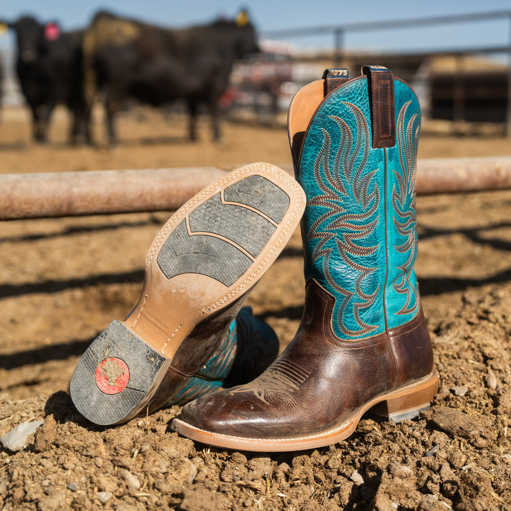 A pair of cowboy boots placed on dirt in an outdoor ranch setting, with one boot standing upright and the other lying on its side to show the sole. The boots are brown leather with metallic turquoise blue decorative stitching and shaft.
