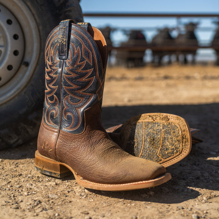 A pair of brown leather cowboy boots with dark blue and tan western embroidery, photographed outdoors on a dirt ground near a vehicle tire, with the boot sole facing upward.