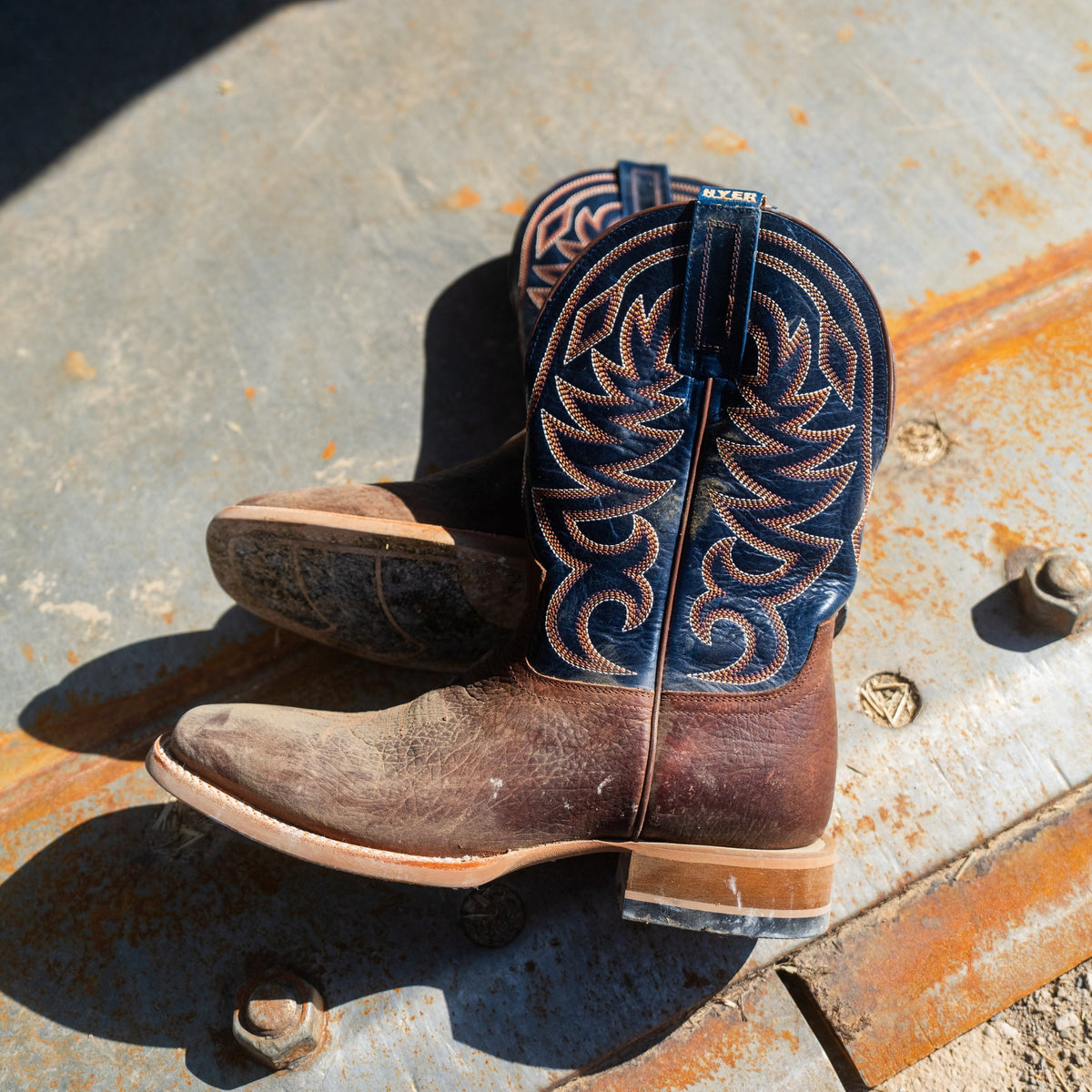 A pair of western cowboy boots resting on a rusty metal surface outdoors. The boots feature dark brown leather bodies with intricate dark blue and tan embroidery on the shafts.