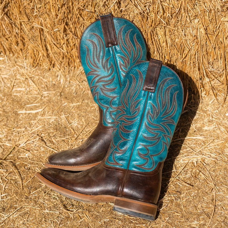 A pair of cowboy boots with dark brown leather and metallic turquoise blue embroidered shafts, displayed on a bed of straw.