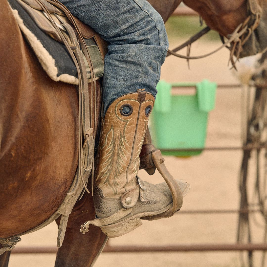 A close-up of a person riding a brown horse, wearing a tan western boots with decorative stitching and blue jeans. The boot is shown in use on the horse's stirrup, with a green bucket and fencing in the background.