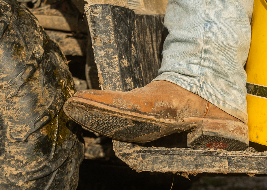 A close-up of a person's foot wearing a dusty brown leather cowboy boot, stepping onto a muddy tractor or farm machine step, with blue jeans and a large mud-covered tire visible.