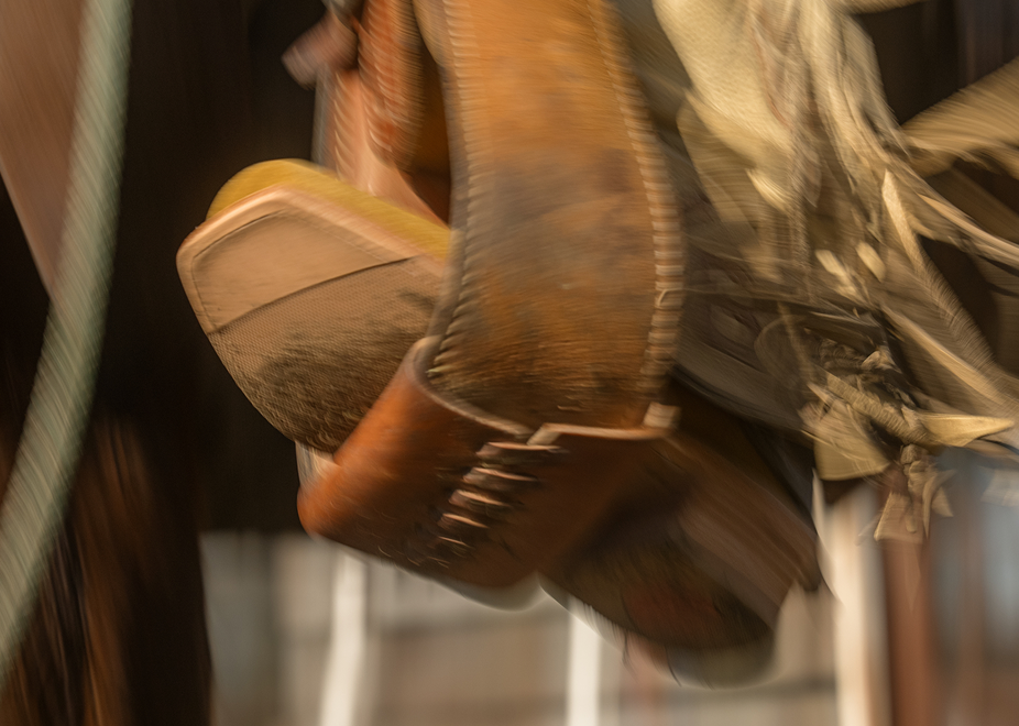 A close-up, motion-blurred shot of a brown leather cowboy boot , held against a dark background with some light-colored fringe or fabric visible on the right.