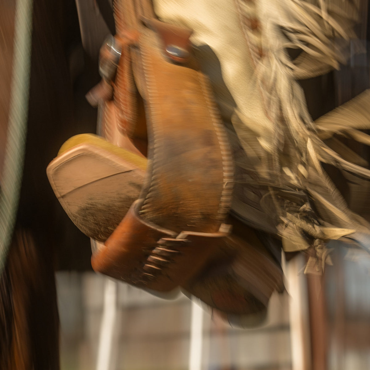 A close-up, motion-blurred shot of brown leather chaps, with a fringed western hat and boots in the background.