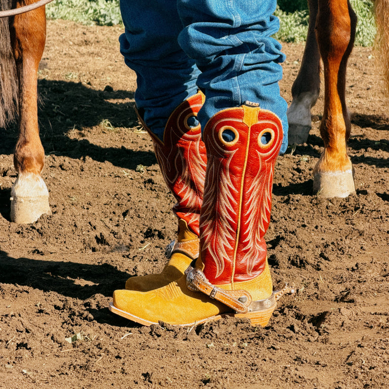 A close-up of a person's legs wearing bright red and tan embroidered cowboy boots with blue jeans, standing in a dirt paddock with parts of two horses visible on both sides.
