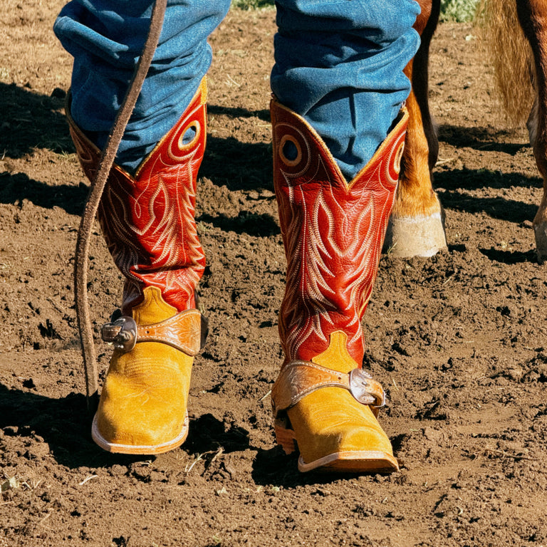 A close-up of a person's legs wearing tan and brown embroidered cowboy boots with blue jeans, standing on dusty brown dirt next to a horse.