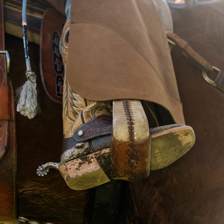 A close-up of a cowboy boot with ornate floral embroidery and a silver concho, tucked into a brown leather chaps leg cover, with a horse's leg visible in the background.