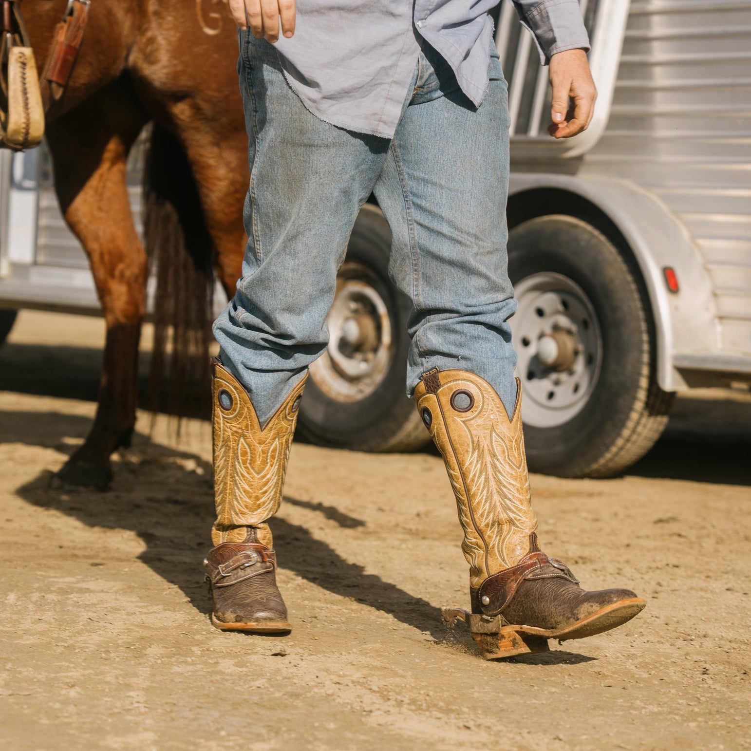 A person wearing tall western-style boots with decorative stitching and dark brown buckles, paired with blue jeans, standing on a dirt surface near a horse and a white trailer.