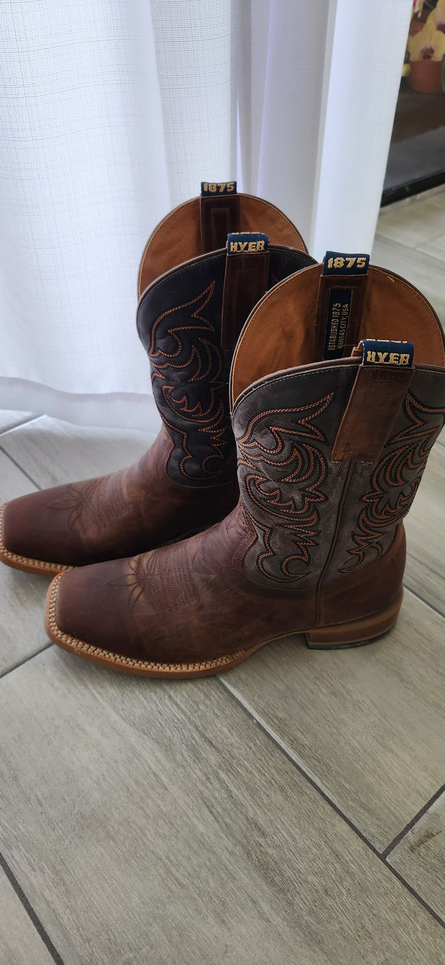 A pair of brown leather cowboy boots with dark brown tooled shafts and light tan stitching, standing on a light gray tile floor in front of a white curtain.