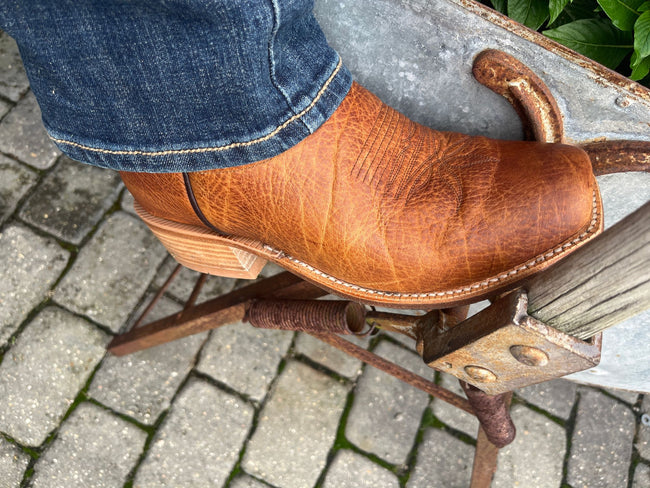 A close-up of a person's foot wearing a brown leather cowboy boot, resting on a rustic metal stand over a paved stone ground.