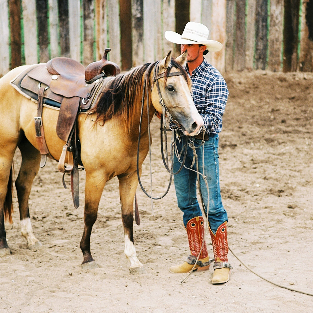 A man in a white cowboy hat, blue plaid shirt, jeans, and reddish brown cowboy boots stands beside a tan horse with a brown western saddle in a dusty outdoor arena.