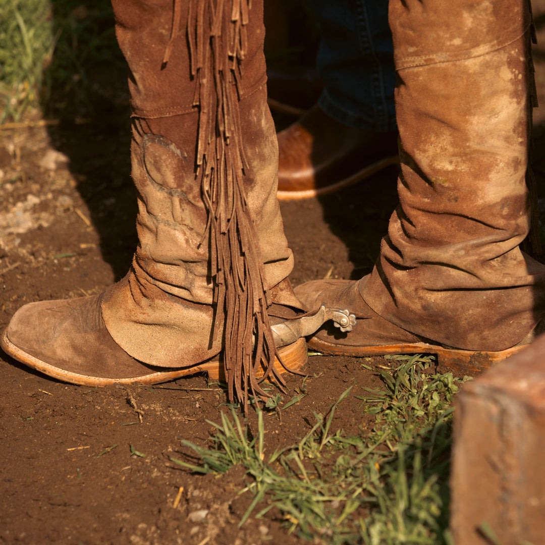 Close-up of a person wearing distressed brown suede fringe chaps standing outdoors on dirt ground with grass.