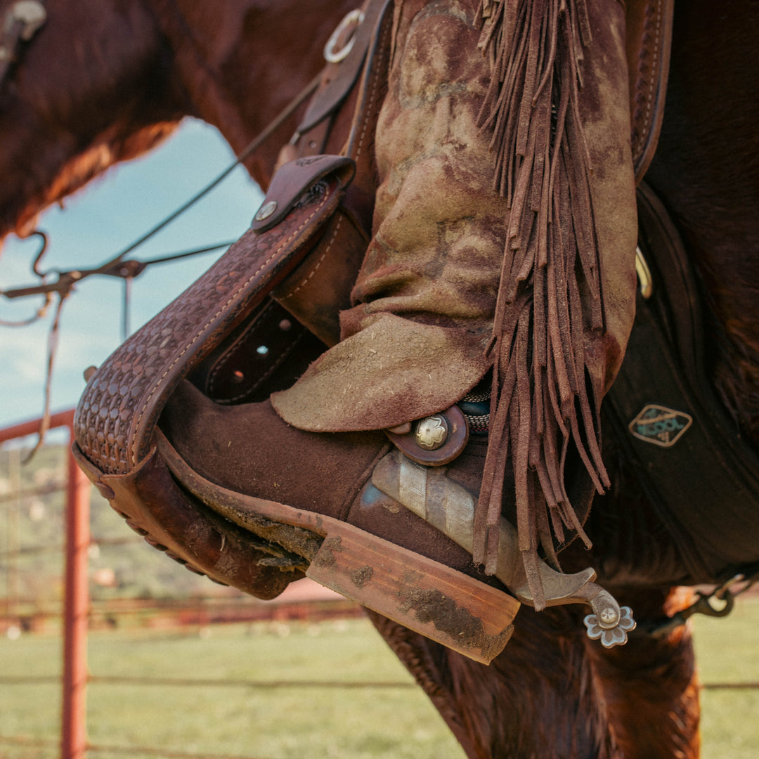 A close-up of a rider's leg in chaps and a brown riding boot, seated on a horse with a leather bridle and spur visible against an outdoor fence background.