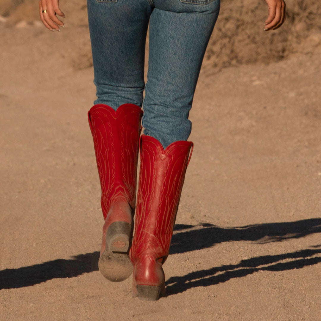 Red cowboy boots worn with blue jeans on a dirt ground.