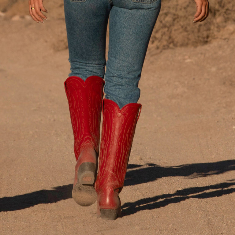 Red cowboy boots worn with blue jeans on a dirt ground.