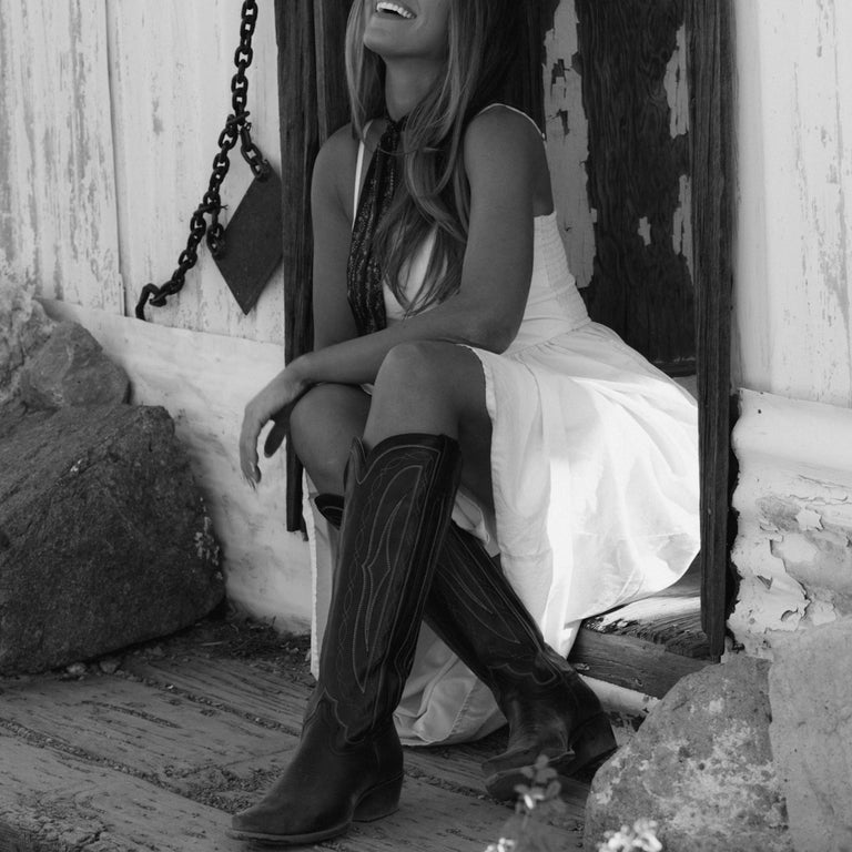 Woman in a white dress and black cowboy boots sitting on a rustic wooden bench.