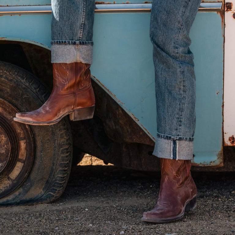 Person wearing brown cowboy boots and blue jeans standing next to a vintage truck.