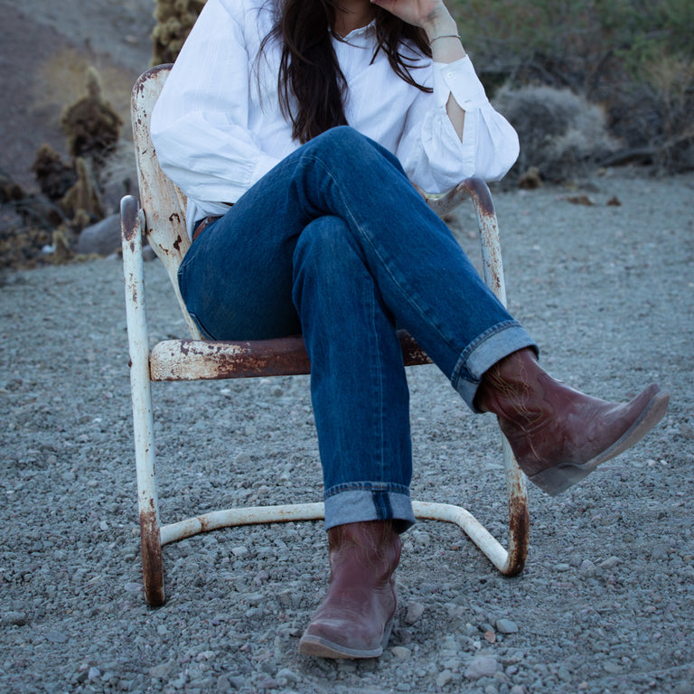 Person sitting on a rusted chair in a desert-like setting wearing a white shirt, blue jeans, and brown boots.
