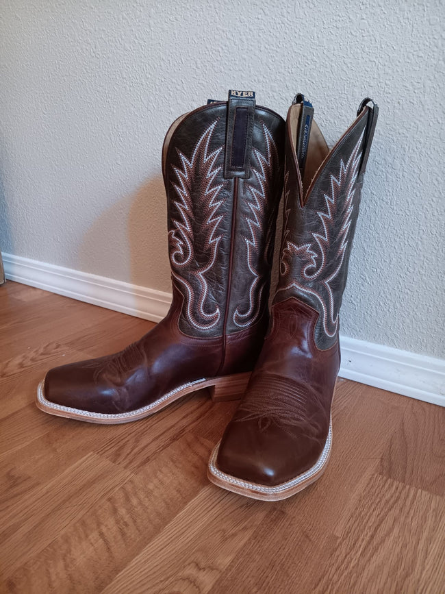 A pair of dark brown leather cowboy boots with white embroidered stitching, standing on a light wood floor against a textured off-white wall.
