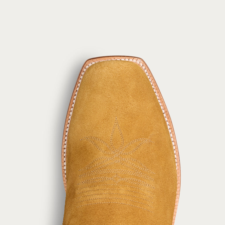 Close-up top view of a tan suede boot toe with decorative stitching and a light pinkish-beige welt edge, shown on a light gray background.