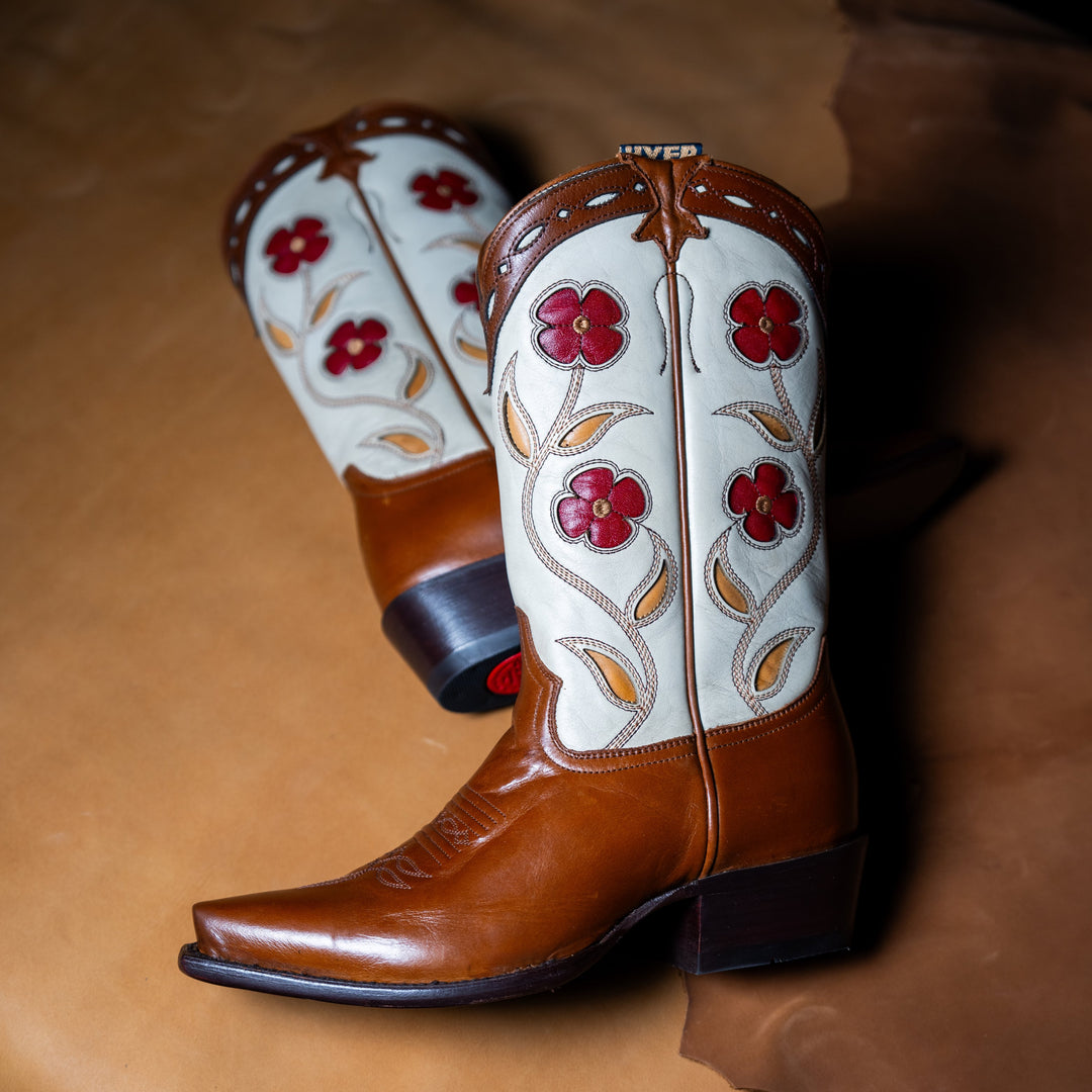 A pair of brown leather cowboy boots with white embroidered shafts featuring red flowers and tan leaves, displayed on a brown leather background.