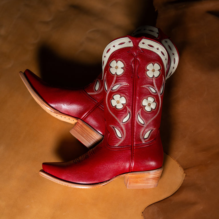 A pair of red leather cowboy boots with white floral embroidery and white trim, displayed on a brown leather background.