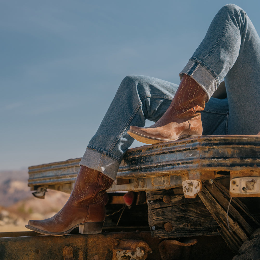Person wearing brown cowboy boots and blue jeans sitting on a rustic surface with a clear sky.