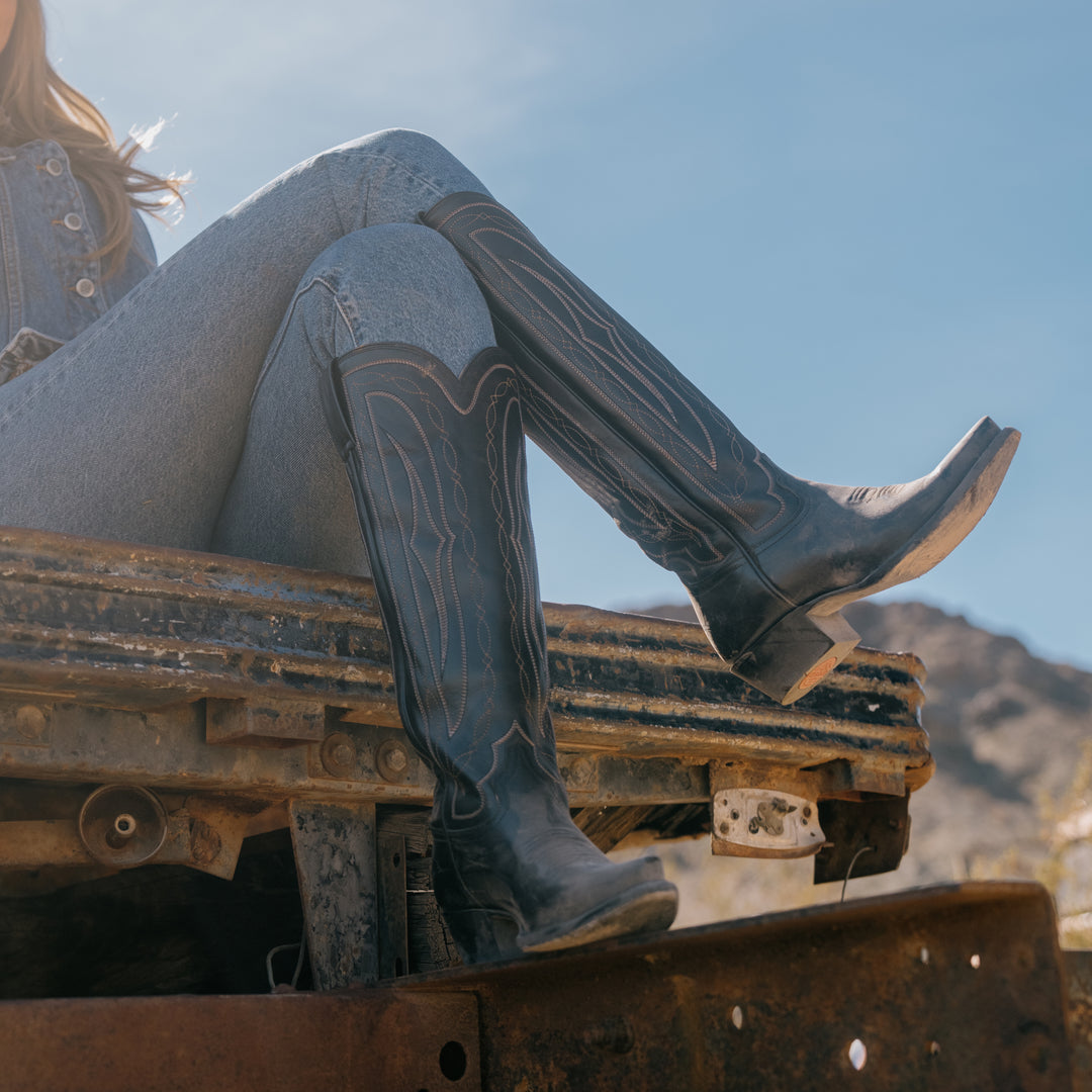 Person wearing cowboy boots sitting on a rusted metal surface with a mountainous background