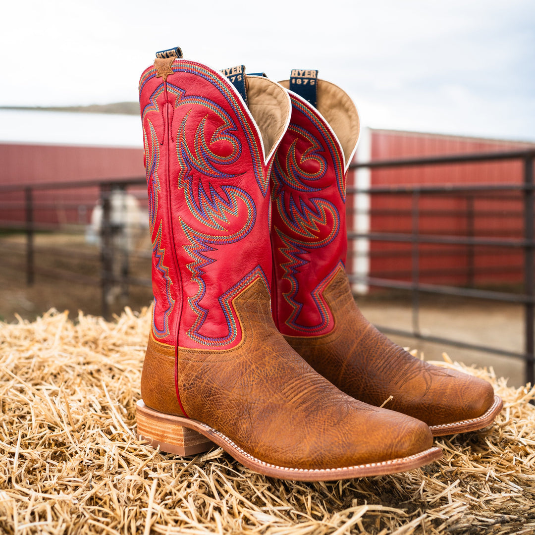 Pair of red and brown cowboy boots on hay with a barn in the background