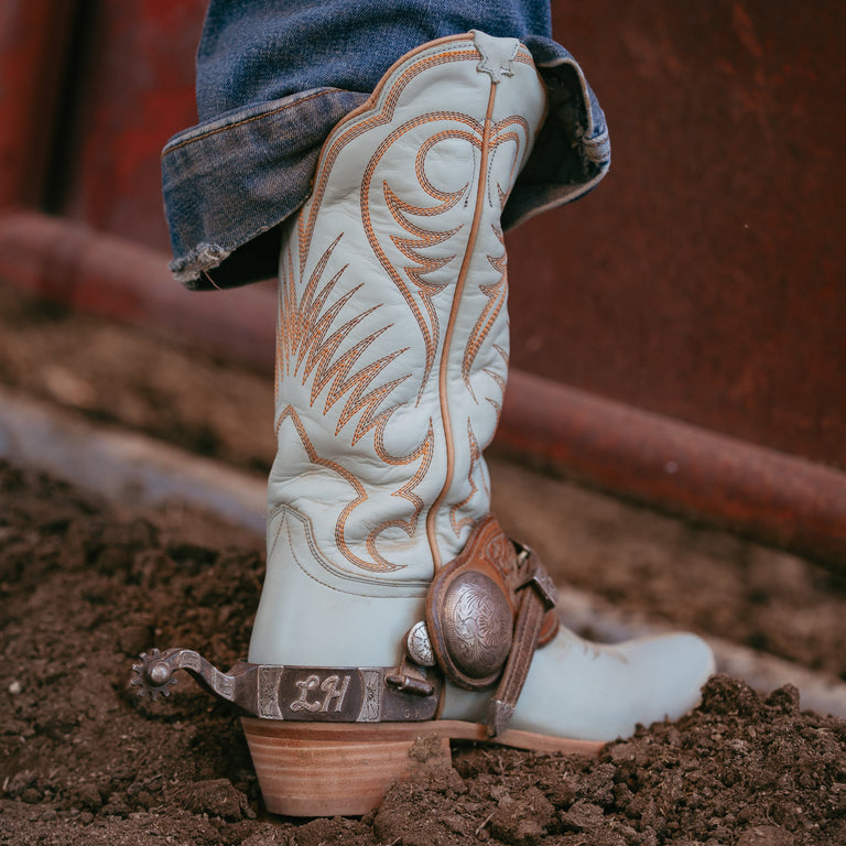 Light blue cowboy boot with brown embroidery and a spur on a dirt ground.