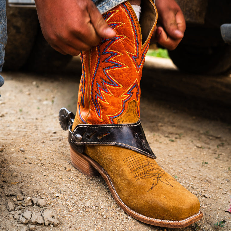 Close-up of a person wearing a tan and orange cowboy boot with intricate leatherwork on a gravel surface.