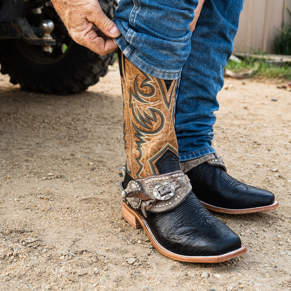 Close-up of a person wearing black and brown cowboy boots on a gravel surface.