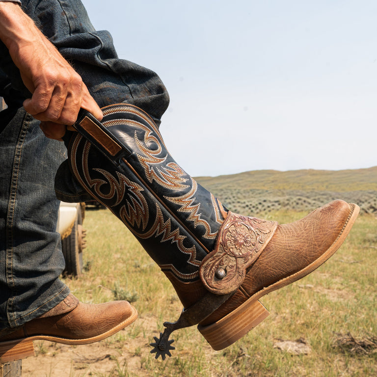Person wearing brown cowboy boots with intricate designs in a natural setting.