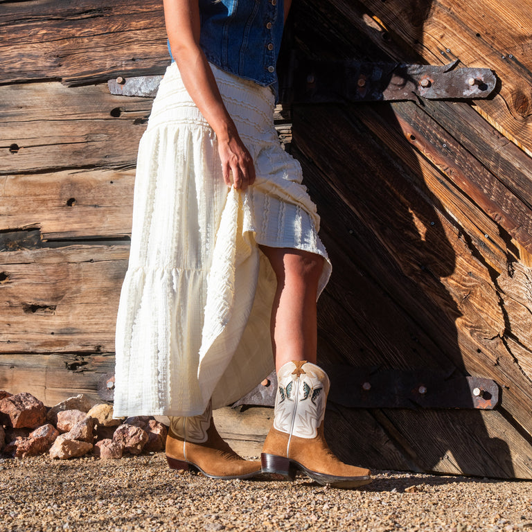 Person wearing white skirt and brown cowboy boots against a wooden wall.