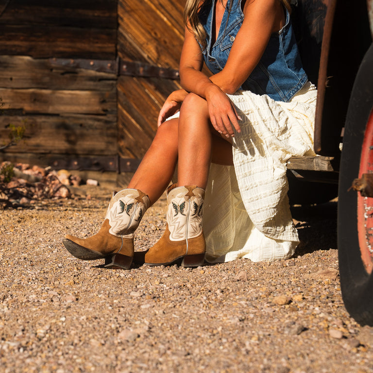Person wearing cowboy boots sitting on a rock with a rustic wooden wall in the background