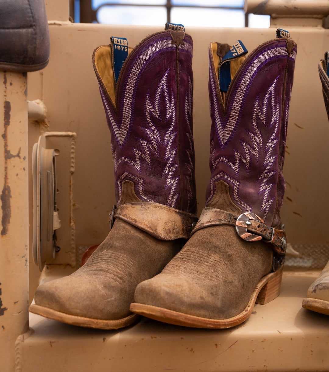 A pair of western cowboy boots with purple embroidered shafts and tan leather feet, resting on a weathered beige wooden surface.