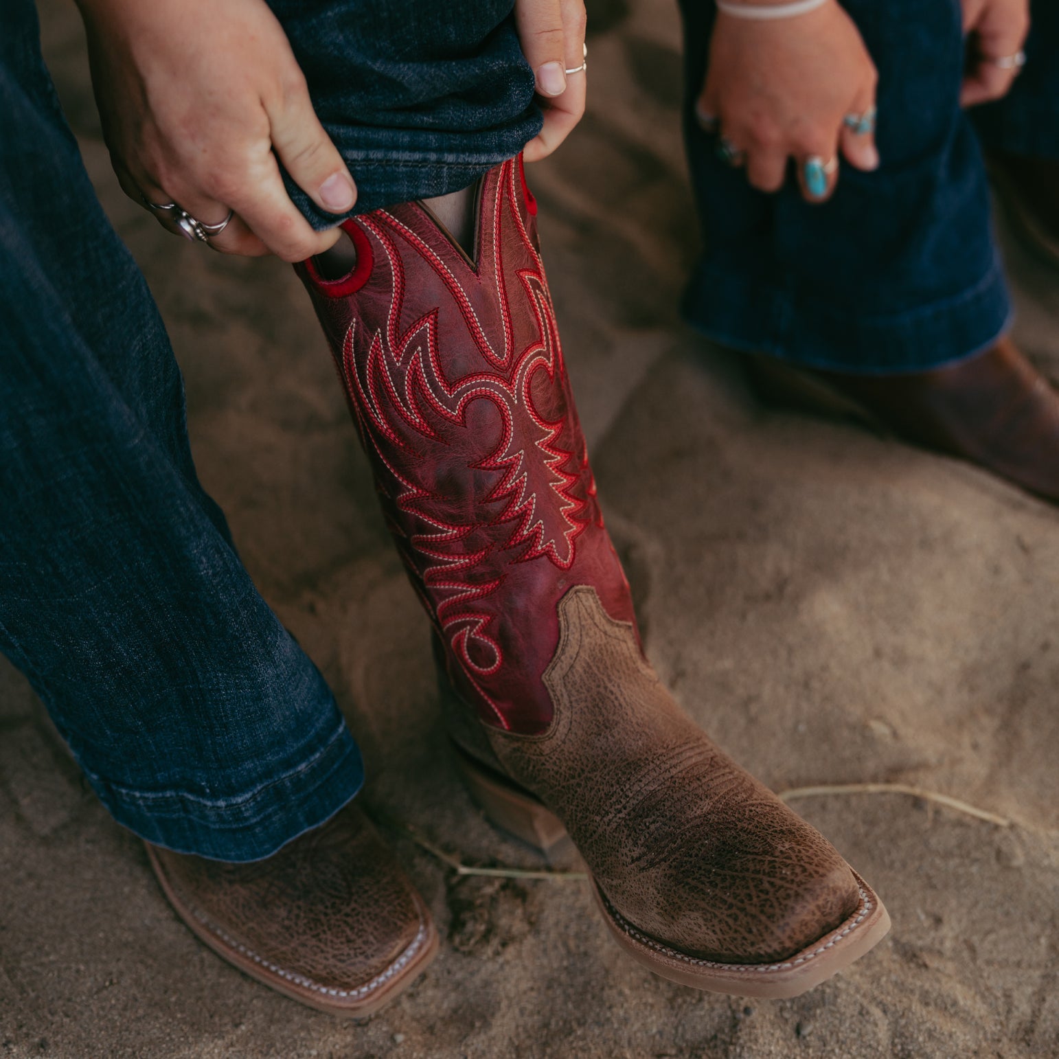 Close-up of a person wearing a brown cowboy boot with red stitching on a sandy ground.