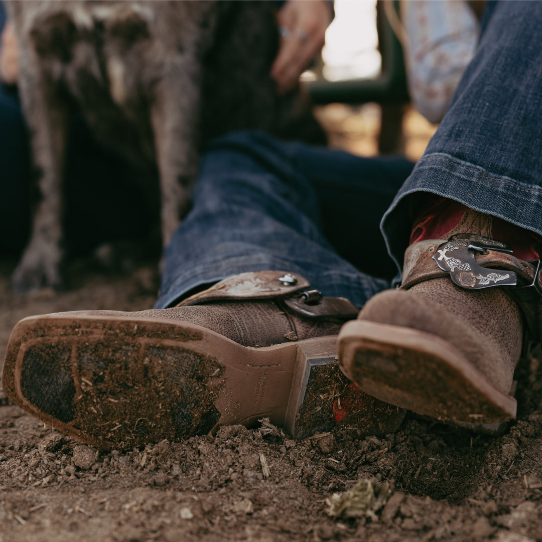 Close-up of brown leather boots with decorative buckles on a dirt ground, with a dog in the background.